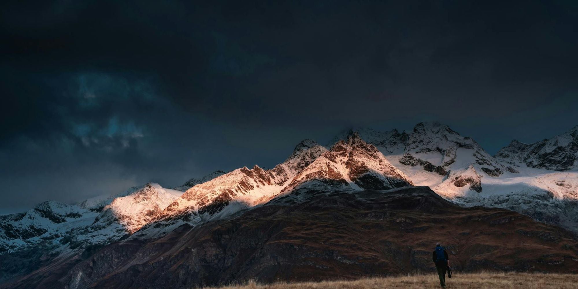 Person standing on a grassy field with snow-capped mountains under a dark, stormy sky.